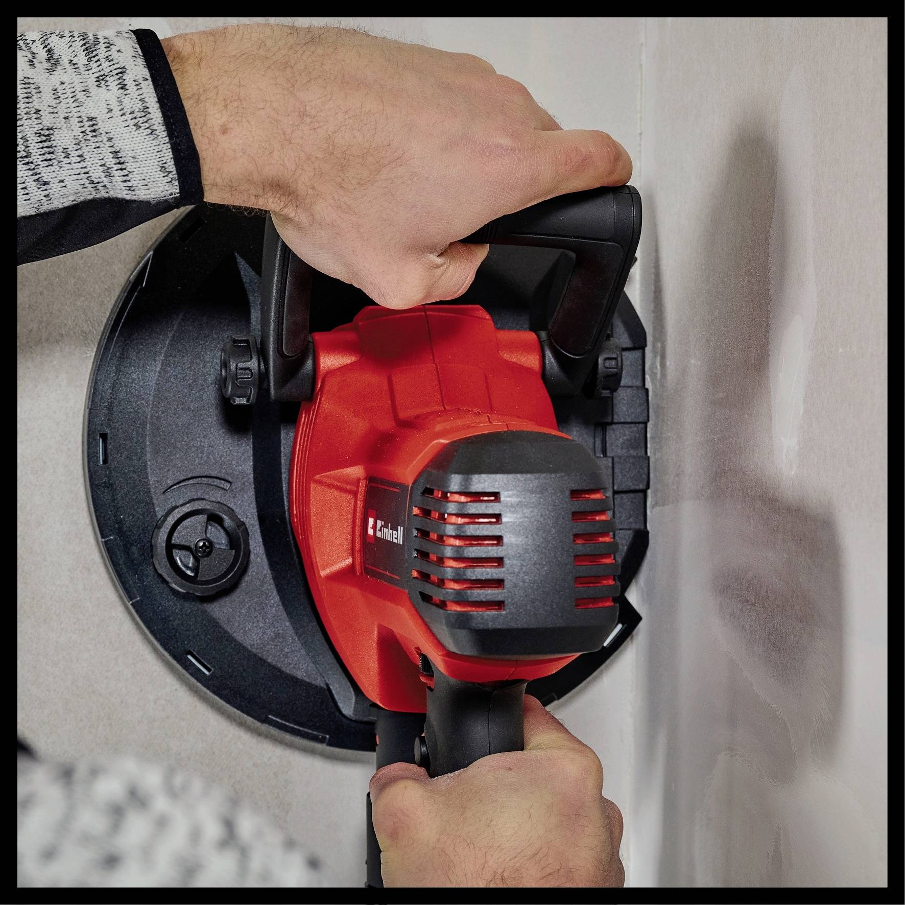 Eine Person hält ein rotes Elektrowerkzeug mit zwei Händen gegen eine Wand. Das Werkzeug ist rund und wird fest an die Wand gedrückt.