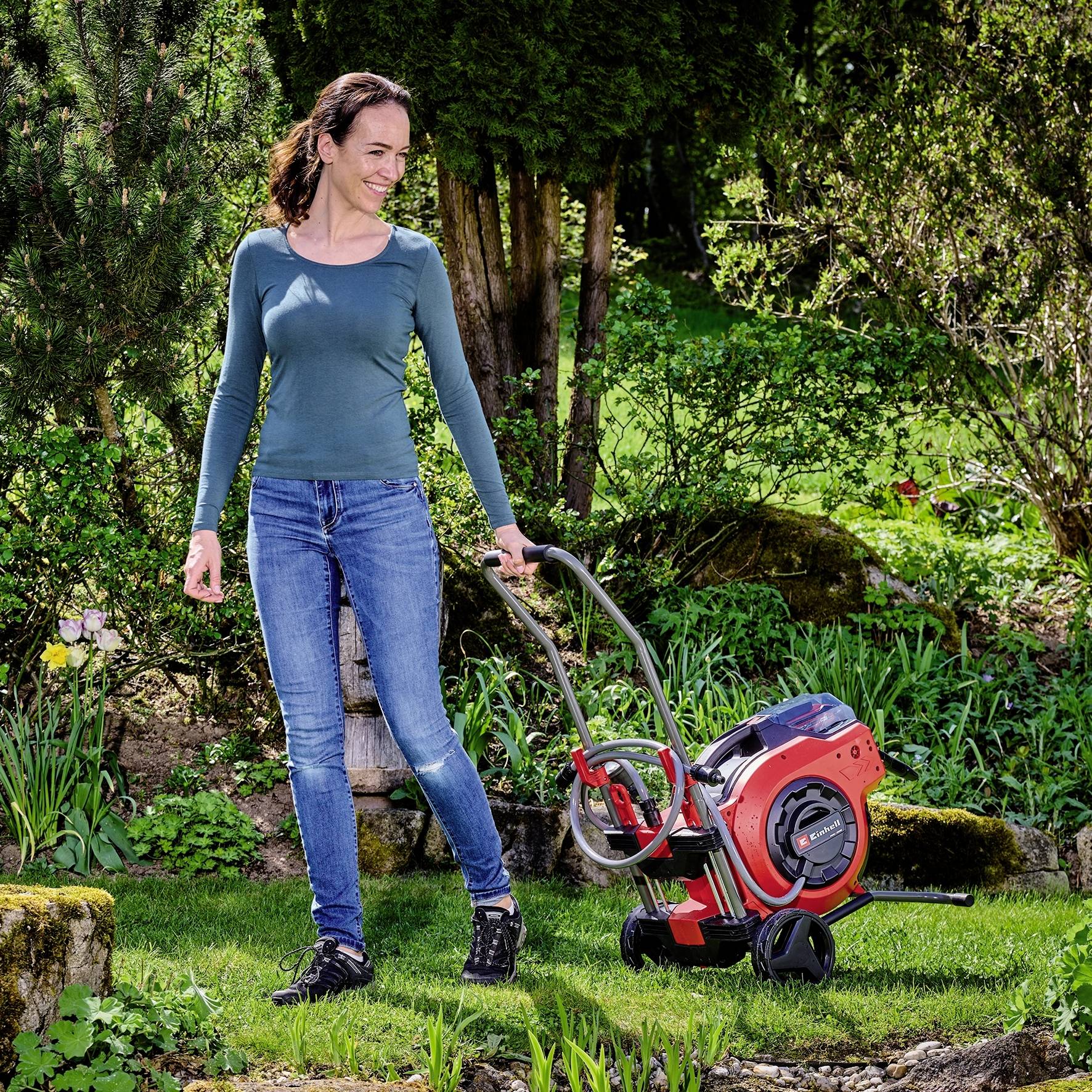 Eine Frau zieht einen roten Rollschneider durch einen gepflegten Garten. Im Hintergrund sind Bäume und Büsche zu sehen.