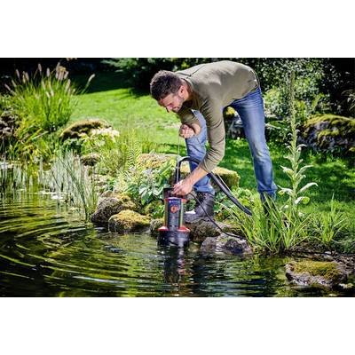 Ein Mann kniet am Ufer eines Teiches und bedient eine Wasserpumpe. Die Umgebung ist grün mit Pflanzen und Moos auf den Steinen.