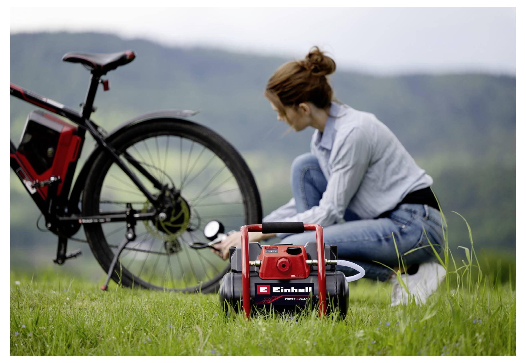 Frau, die neben einem Fahrrad im Gras kniet und mit einem tragbaren Luftkompressor/Reparaturwerkzeug im Freien mit Bergen im Hintergrund arbeitet.