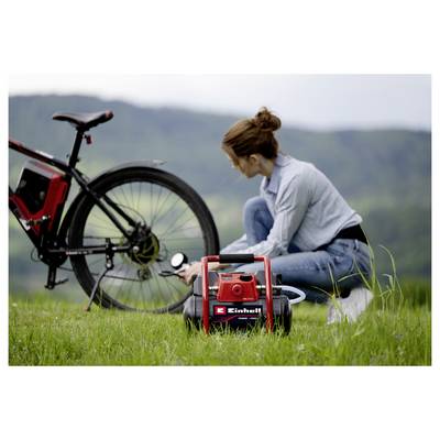 Frau, die neben einem Fahrrad im Gras kniet und mit einem tragbaren Luftkompressor/Reparaturwerkzeug im Freien mit Bergen im Hintergrund arbeitet.