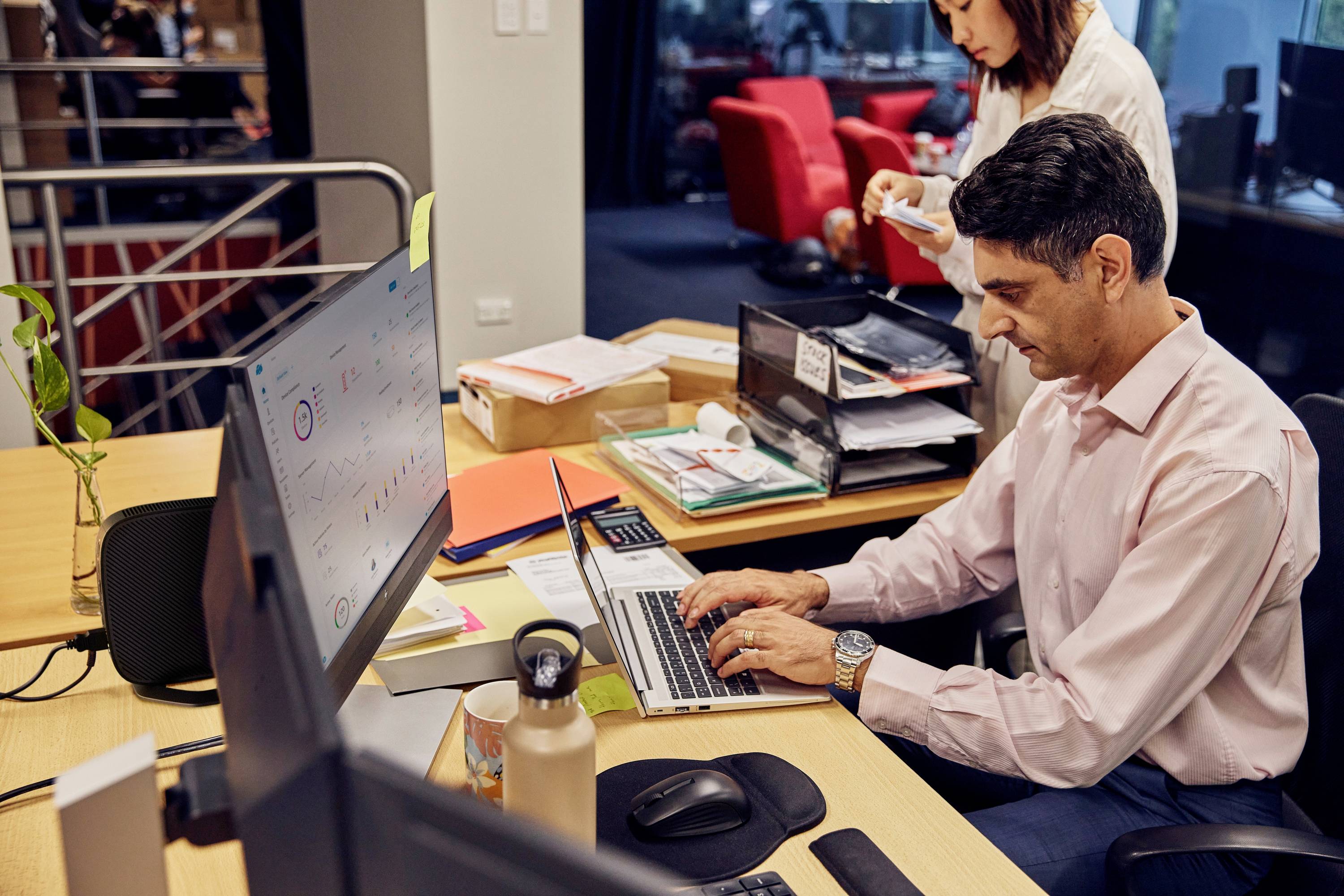Mann auf Arbeitsplatz tippt auf Laptop, Büro mit Papieren und Monitoren, Frau im Hintergrund mit Notizen, geschäftige Szene.