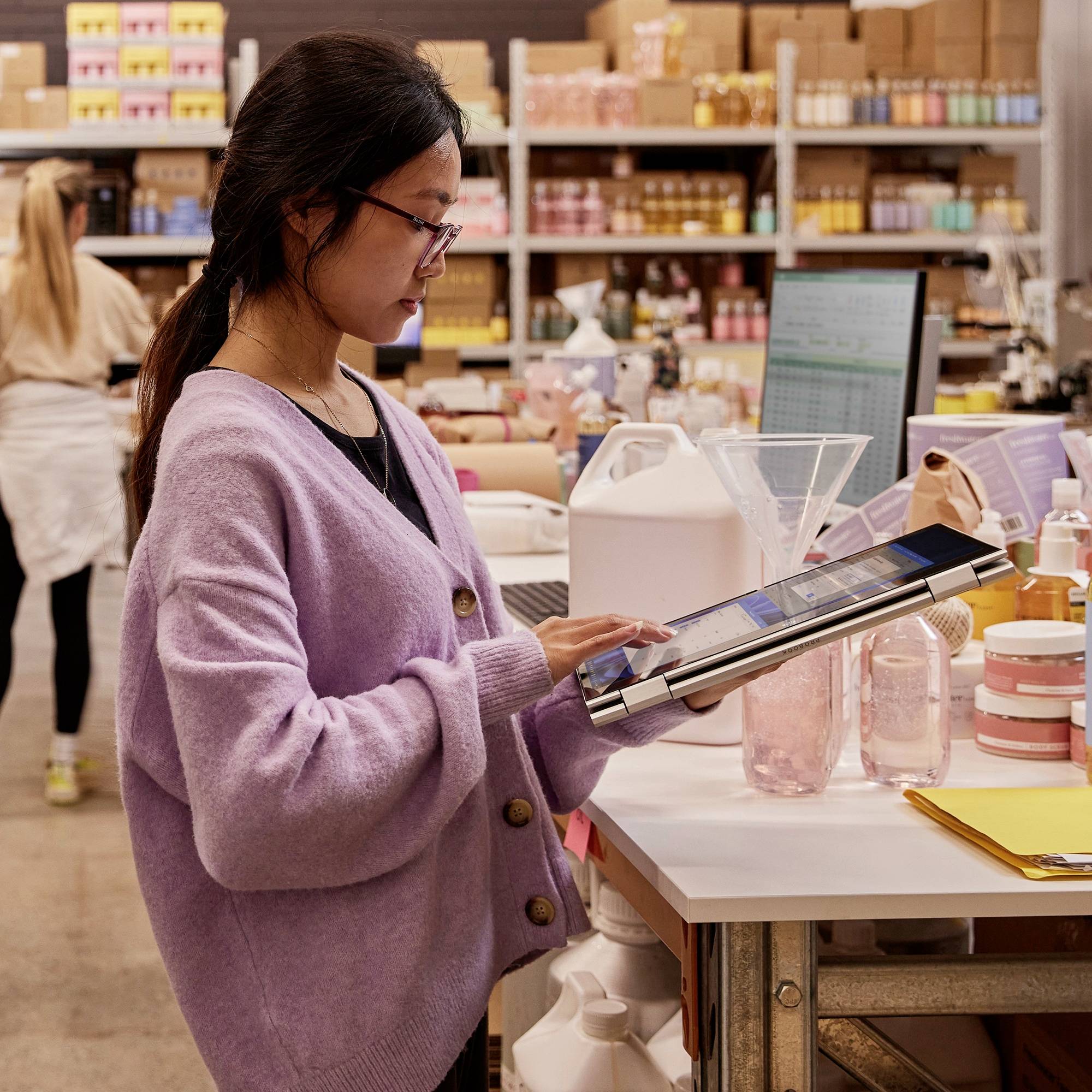 Eine Person in einem Labor hält ein Tablet, umgeben von Flaschen und Geräten. Im Hintergrund sind Regale mit Produkten sichtbar.