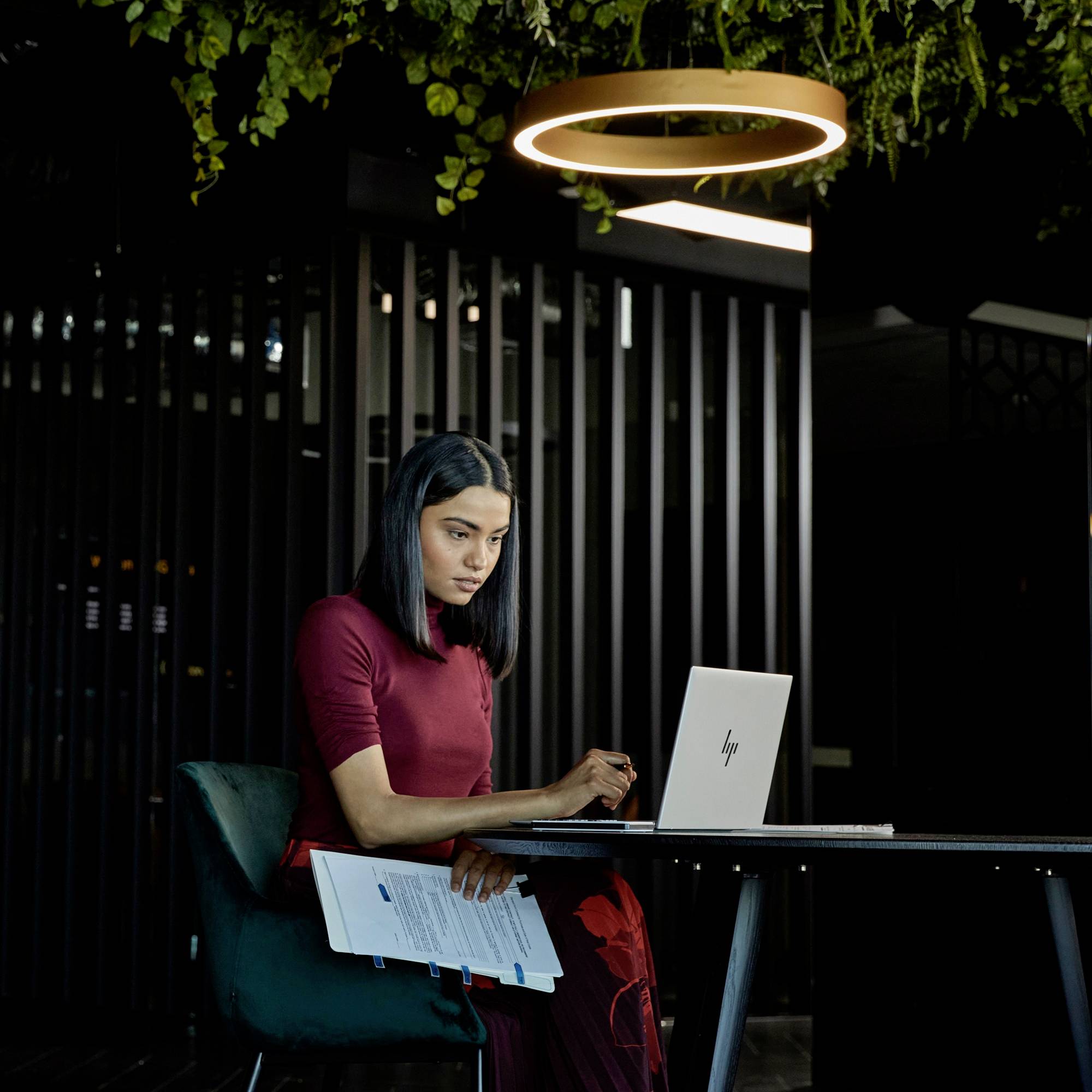 Eine Frau sitzt an einem Tisch und arbeitet konzentriert an einem Laptop. Oberhalb hängt ein beleuchteter Ring mit Pflanzen.