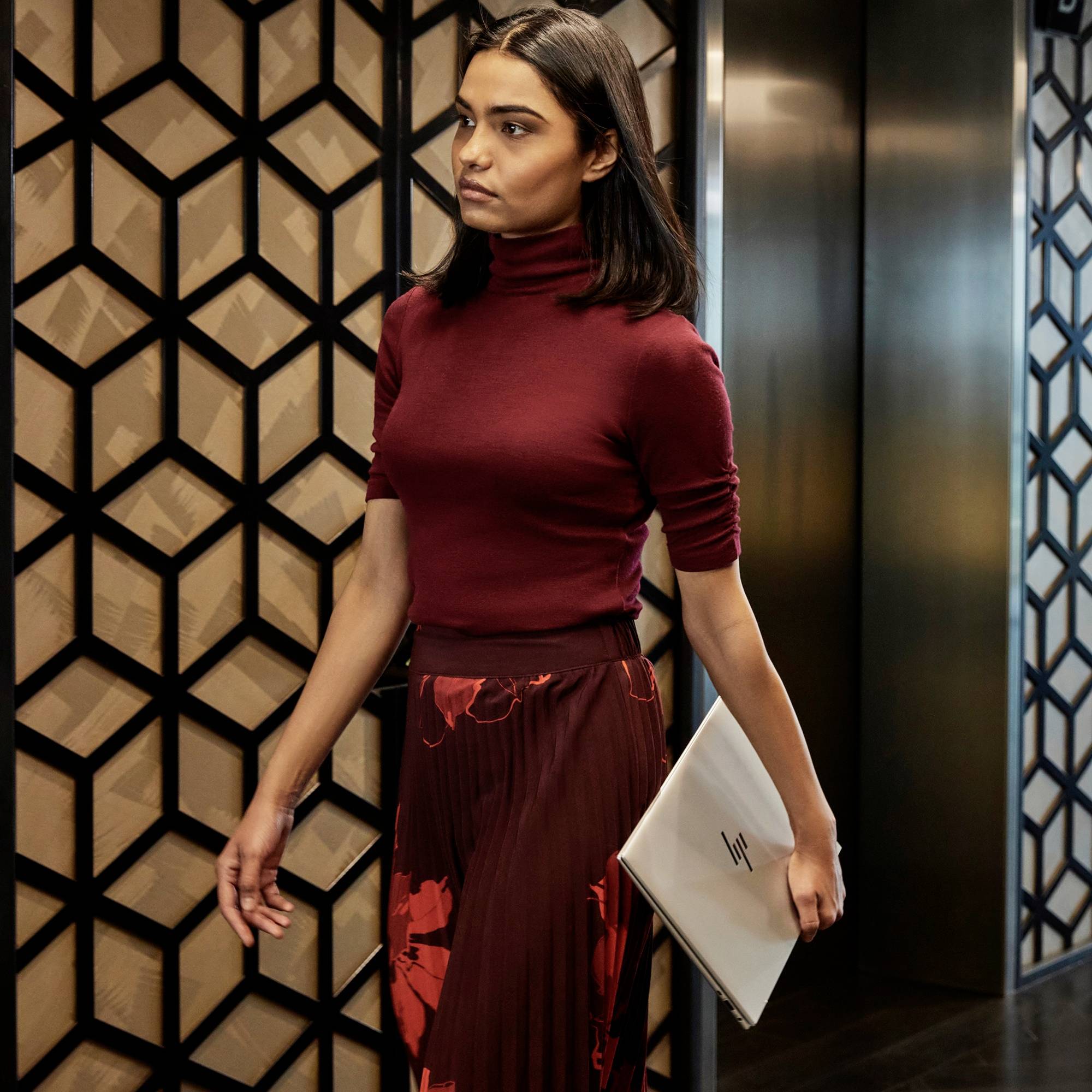 Eine Person in einem eleganten Outfit hält einen Laptop in einem modernen Innenraum mit geometrischen Wänden.
