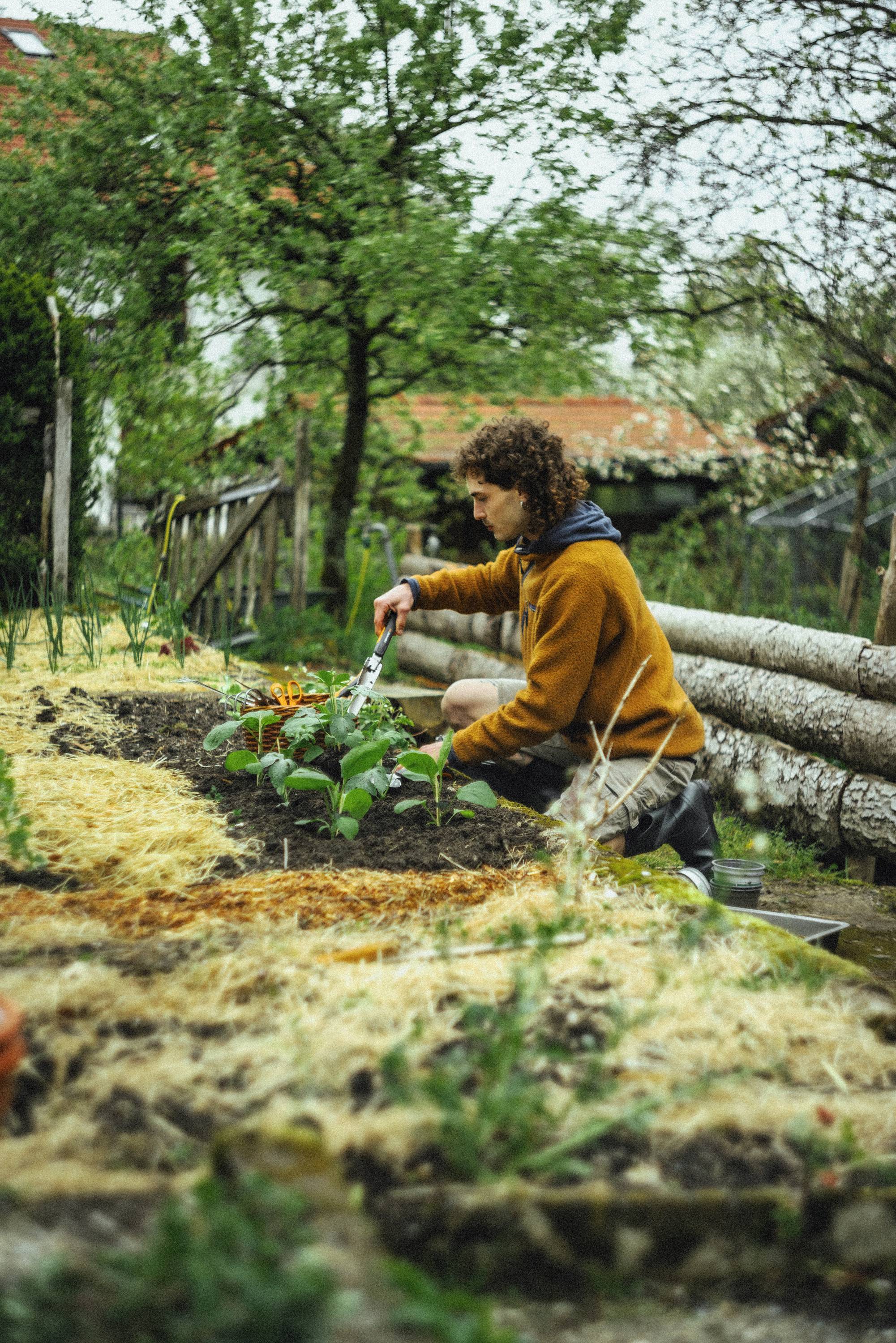 Eine Person in brauner Jacke pflanzt Blumen in einem Garten, umgeben von Bäumen und Holzzäunen.