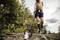 Person greift mit Greifzange eine Plastikflasche in der Natur auf, trägt braunes T-Shirt, schwarze Shorts und Gummistiefel.