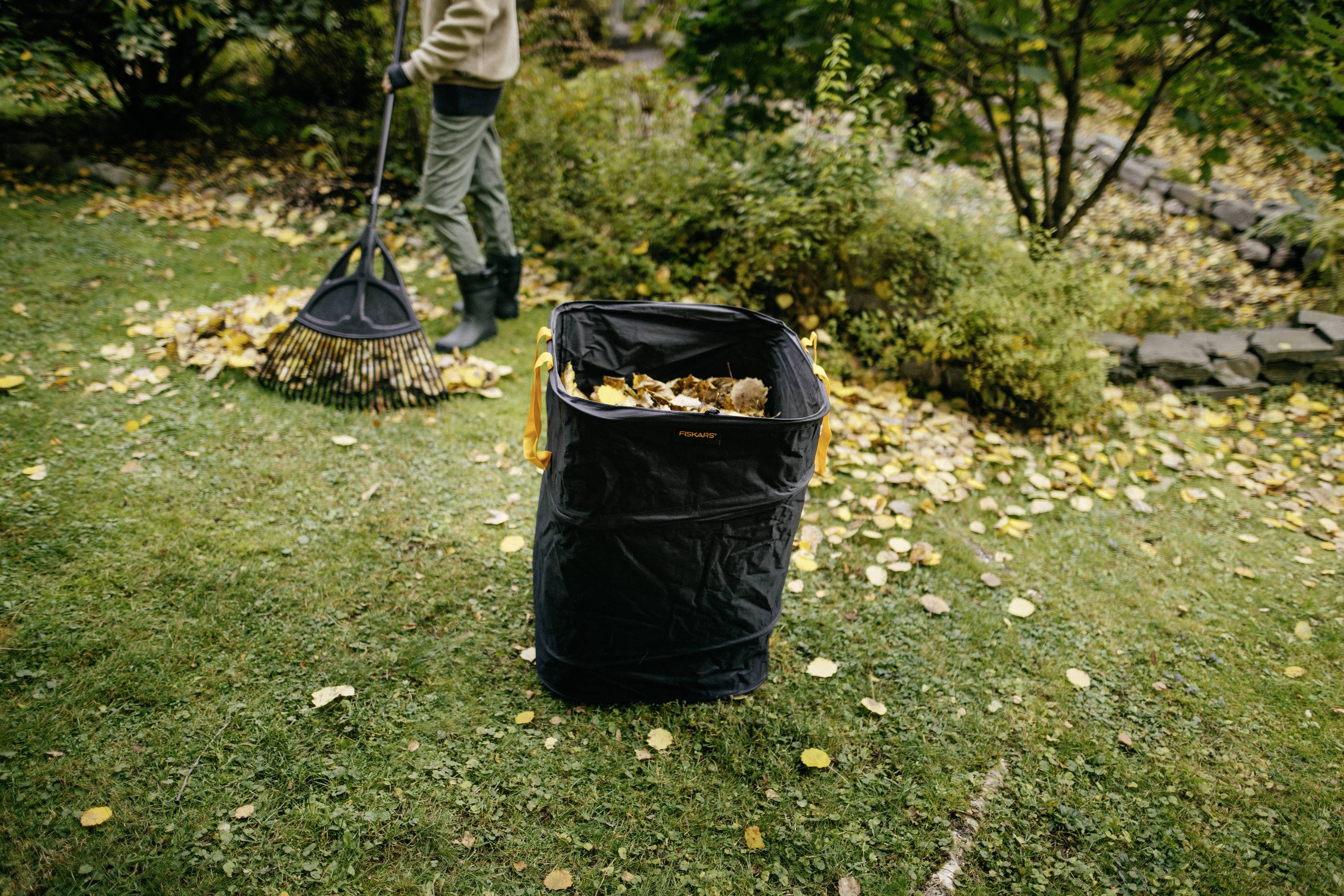 Person harkt Laub im Garten. Ein großer schwarzer Korb im Vordergrund ist mit gelben Blättern gefüllt. Bäume und Sträucher im Hintergrund.