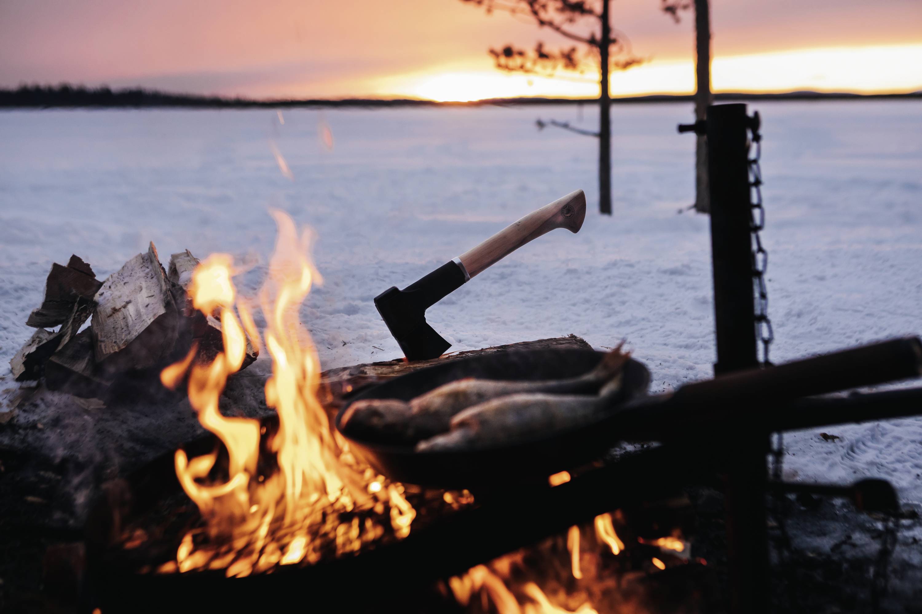 Ein Lagerfeuer brennt vor einer schneebedeckten Landschaft während des Sonnenuntergangs. Eine Axt steckt im Holz, Fische auf dem Grill.