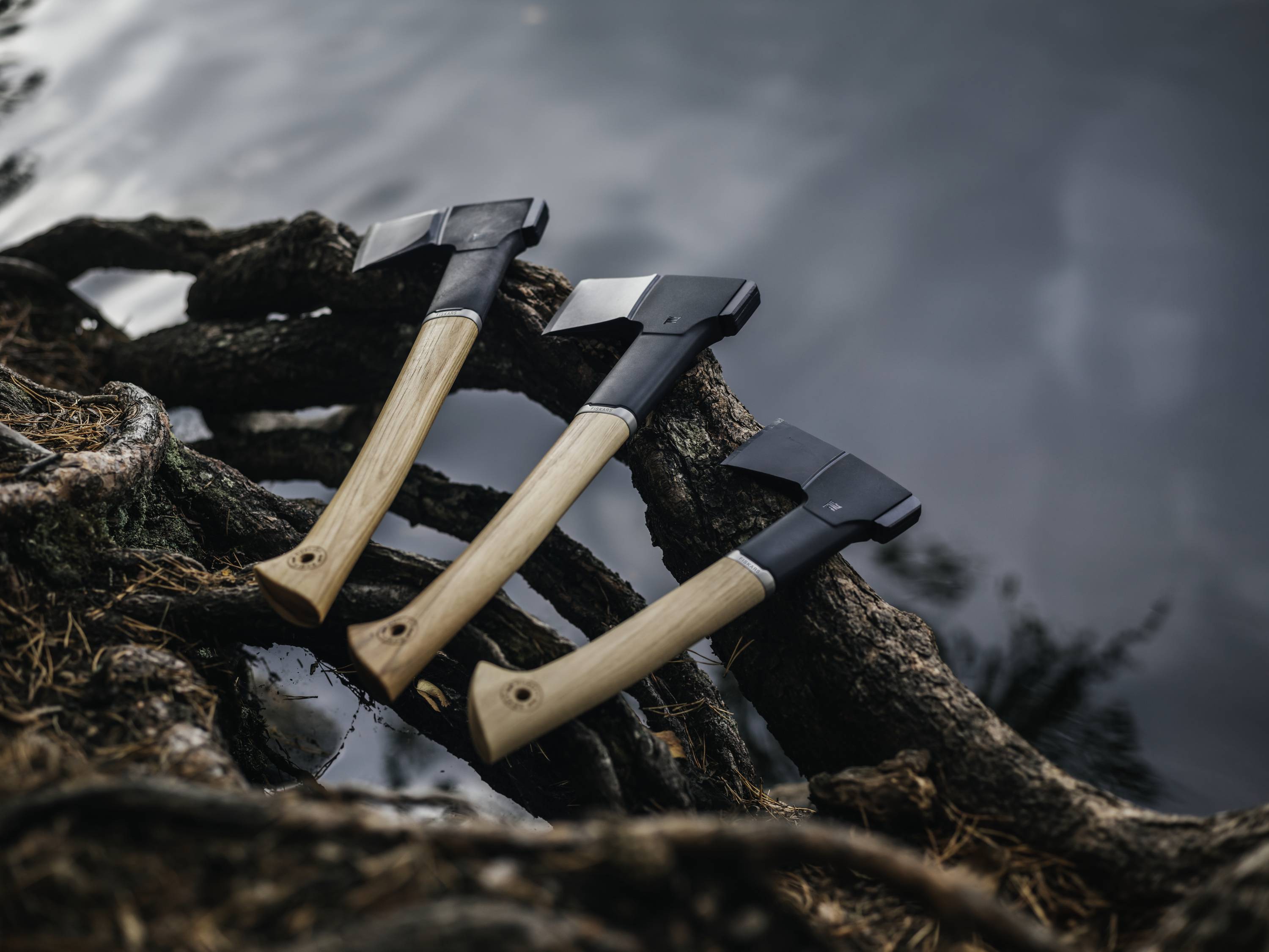Drei Äxte mit Holzgriffen liegen auf dicken, verwurzelten Ästen am Wasser, spiegeln den Übergang von Natur und Handwerk wider.