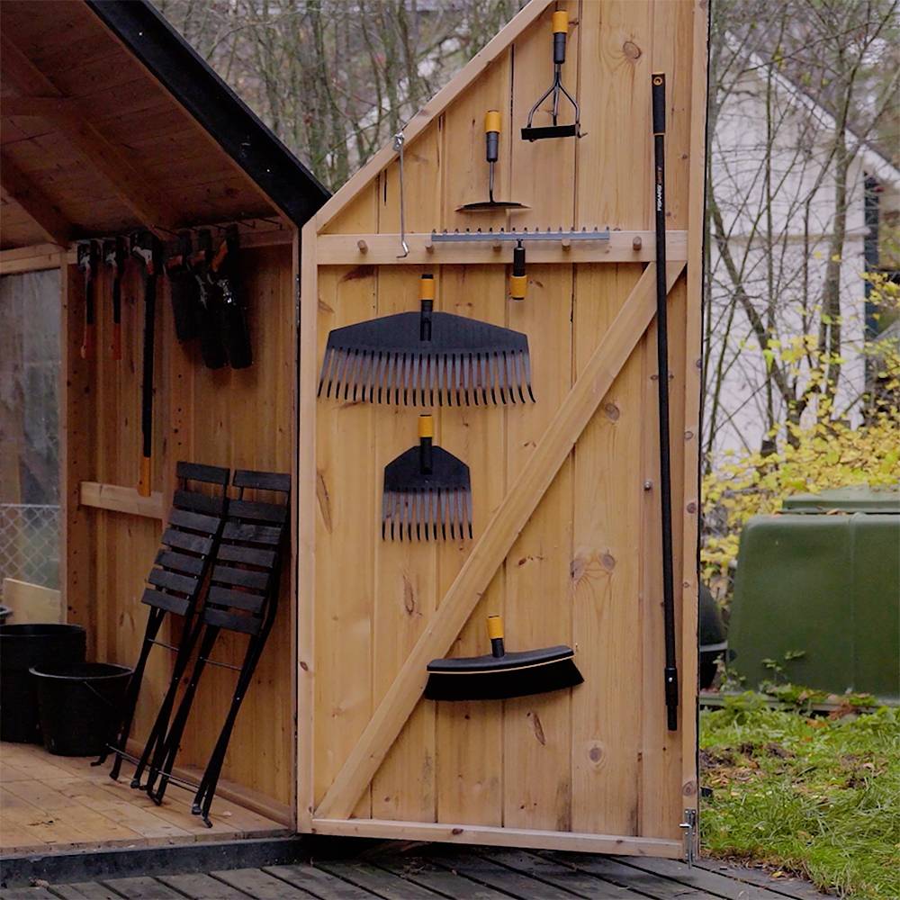 Ein offenes Holzlager mit Gartenwerkzeugen wie Rechen, Schaufeln und Besen, die ordentlich an der Innenwand aufgehängt sind. Im Hintergrund sind Bäume sichtbar.