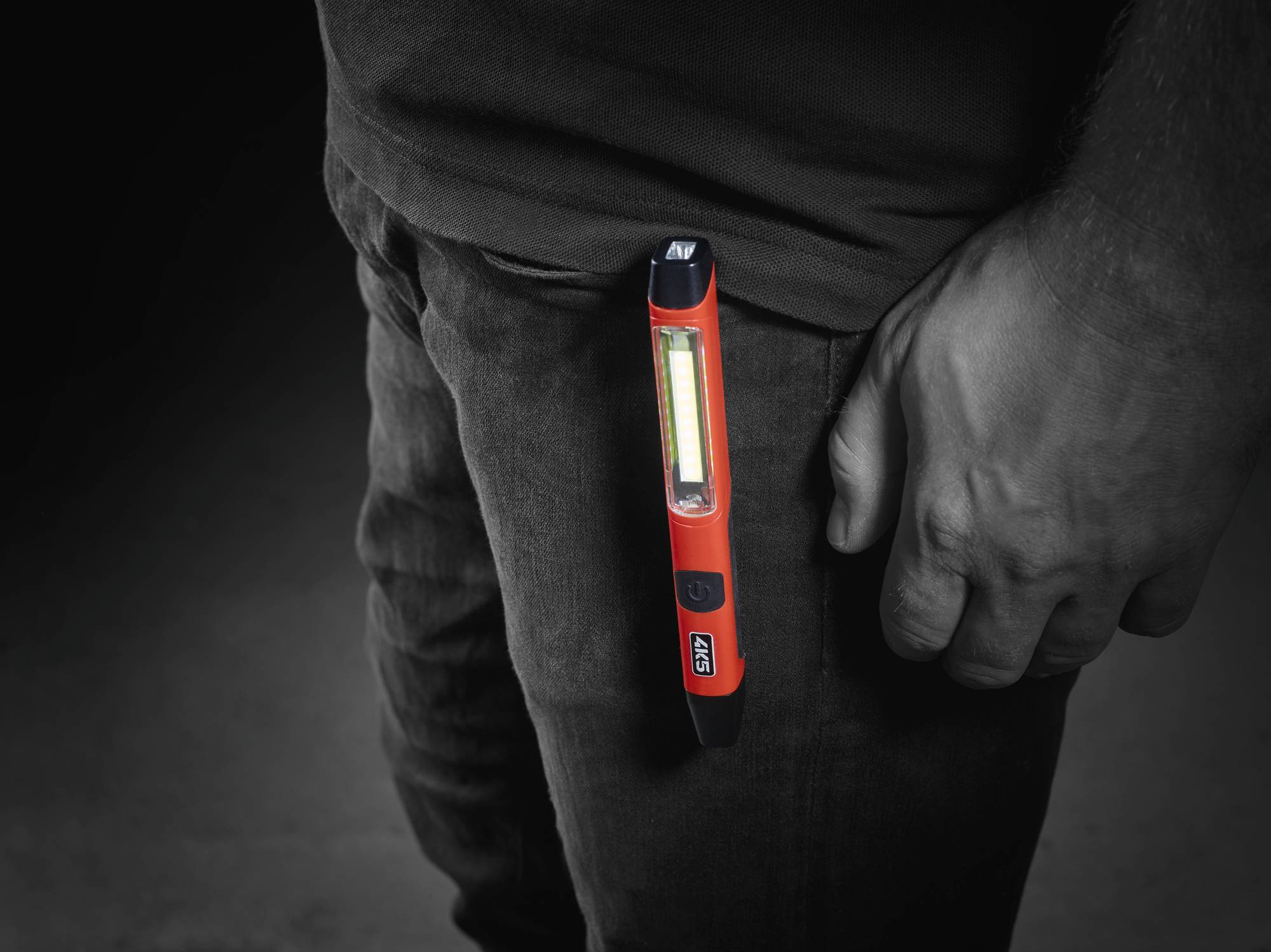 Eine Hand hält eine kleine rote Taschenlampe, die an eine Tasche geklippt ist. Die Lampe leuchtet hell und zeigt nach unten.