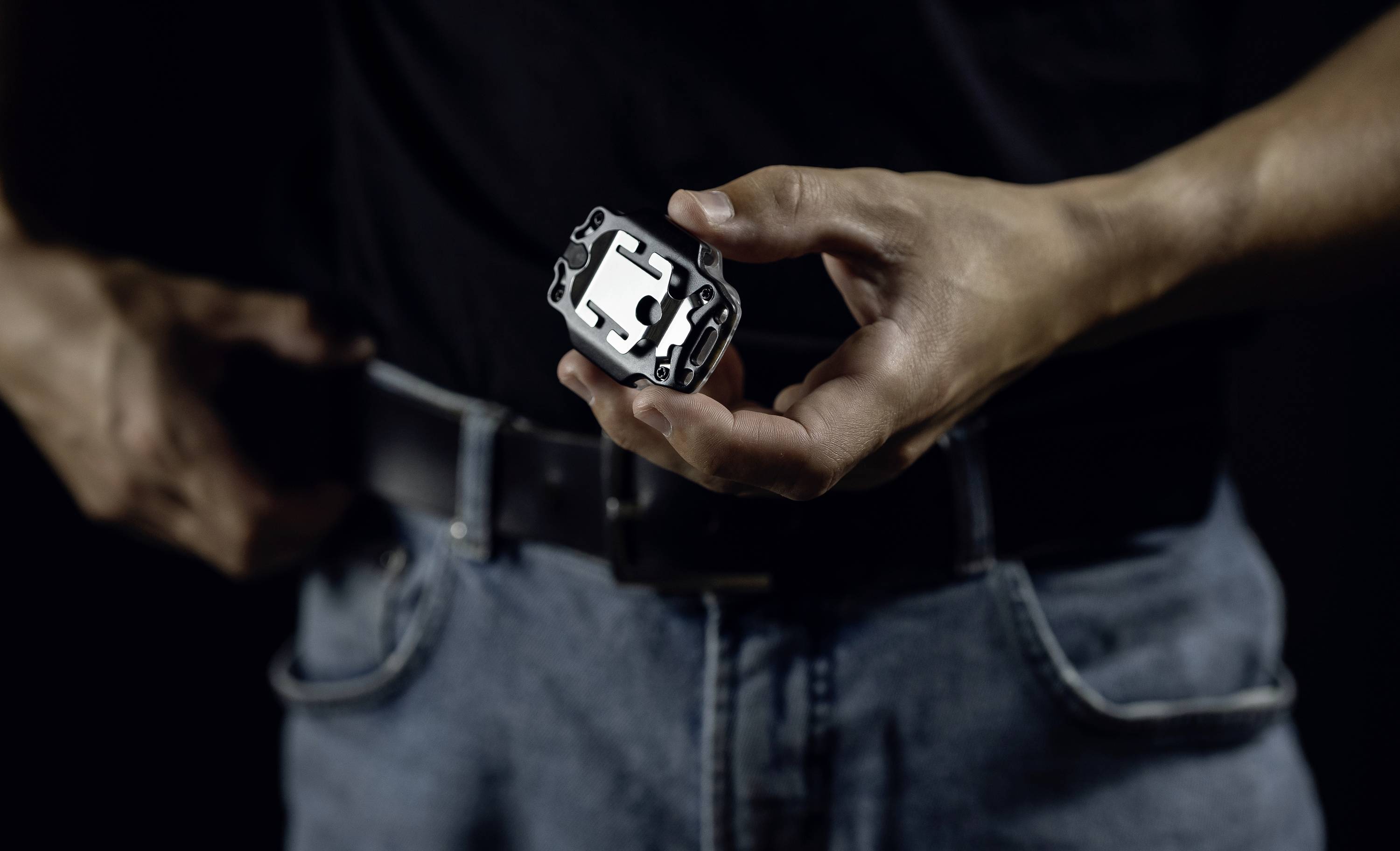 Eine Person hält einen kleinen, rechteckigen Metallgegenstand mit mehreren Aussparungen in der Hand. Die Person trägt Jeans und einen Gürtel.