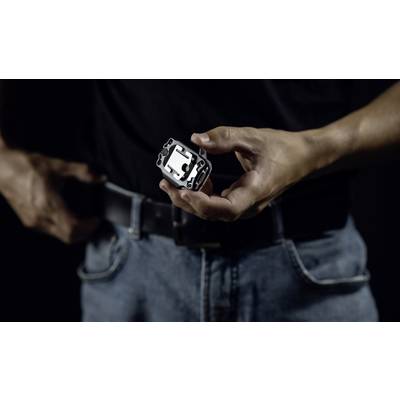Eine Person hält einen kleinen, rechteckigen Metallgegenstand mit mehreren Aussparungen in der Hand. Die Person trägt Jeans und einen Gürtel.