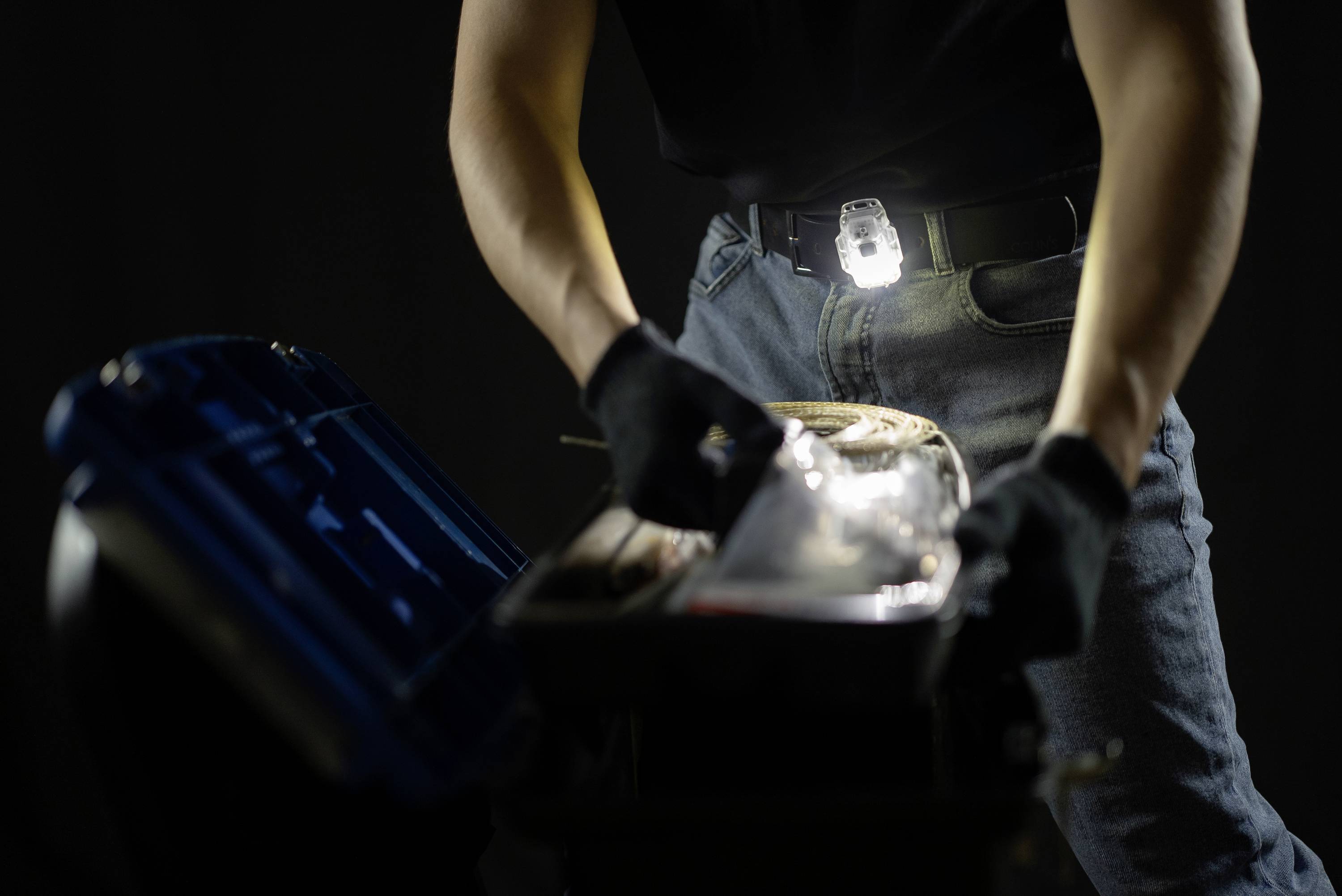 Eine Person arbeitet in einem dunklen Raum mit Taschenlampe und Handschuhen an einer geöffneten Werkzeugkiste.