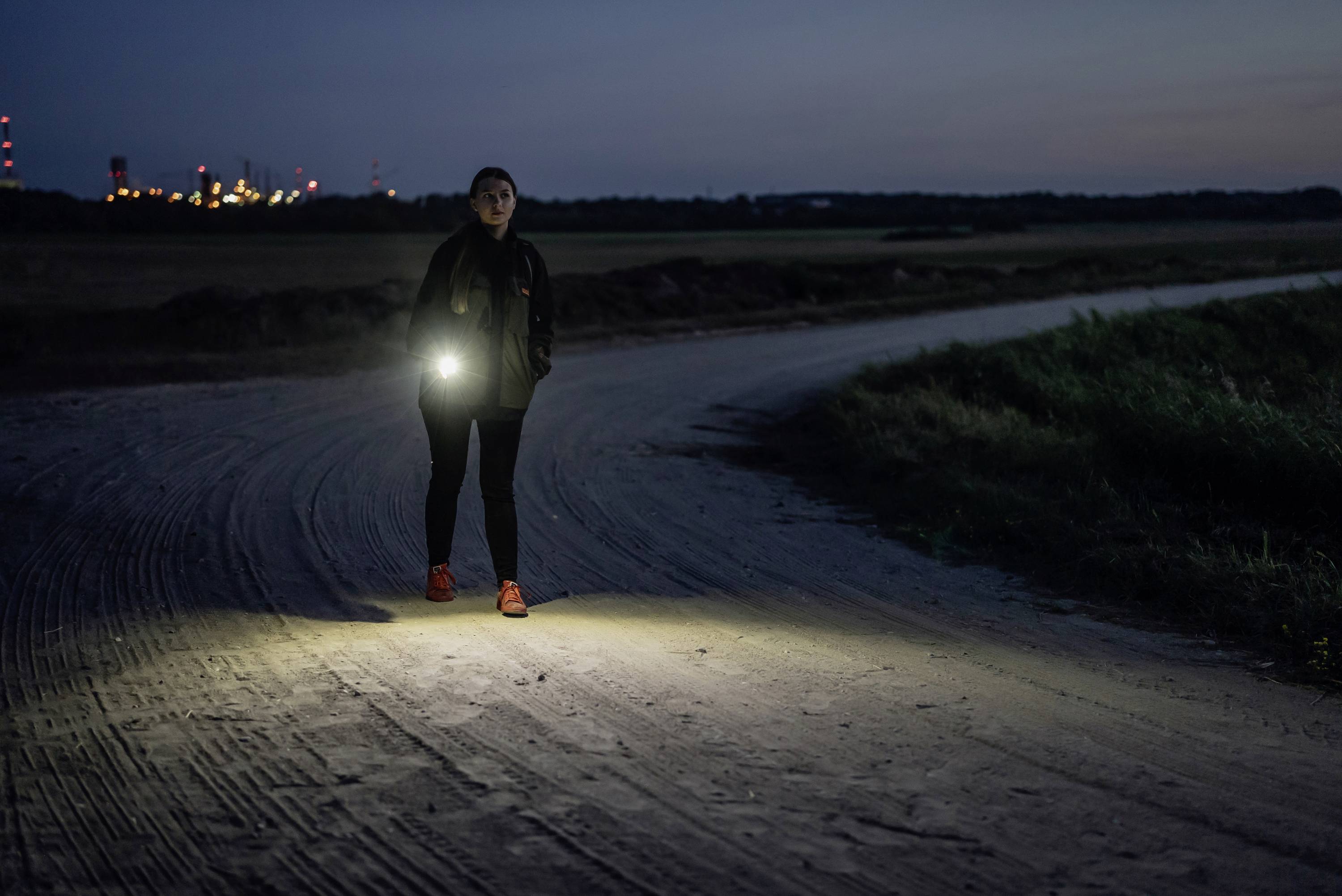 Eine Person geht nachts auf einem Feldweg und trägt eine leuchtende Taschenlampe. Im Hintergrund sind Lichter einer Stadt zu sehen.