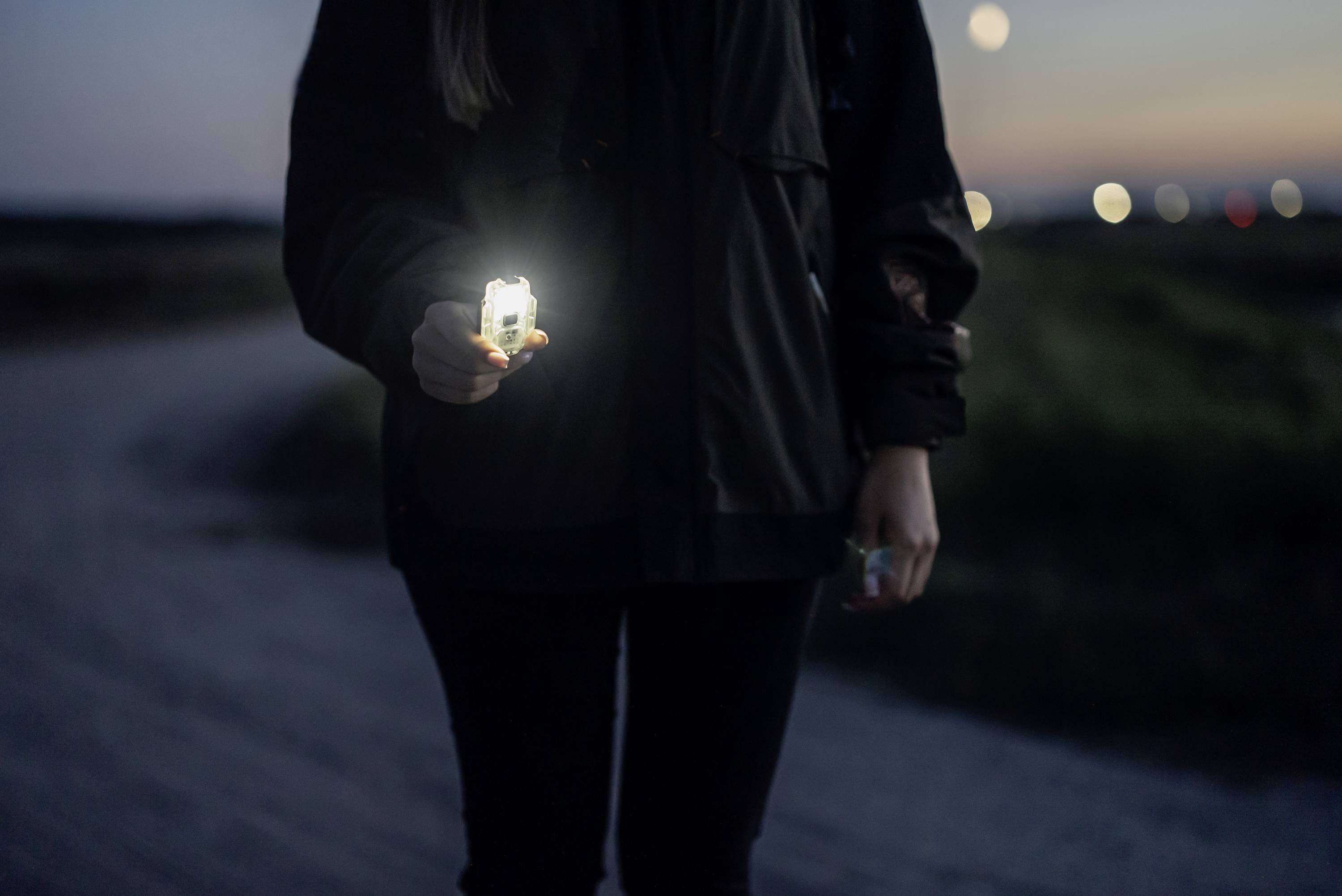 Eine Person hält eine leuchtende Lampe in der Hand. Es ist Abenddämmerung und sie steht auf einem Weg.