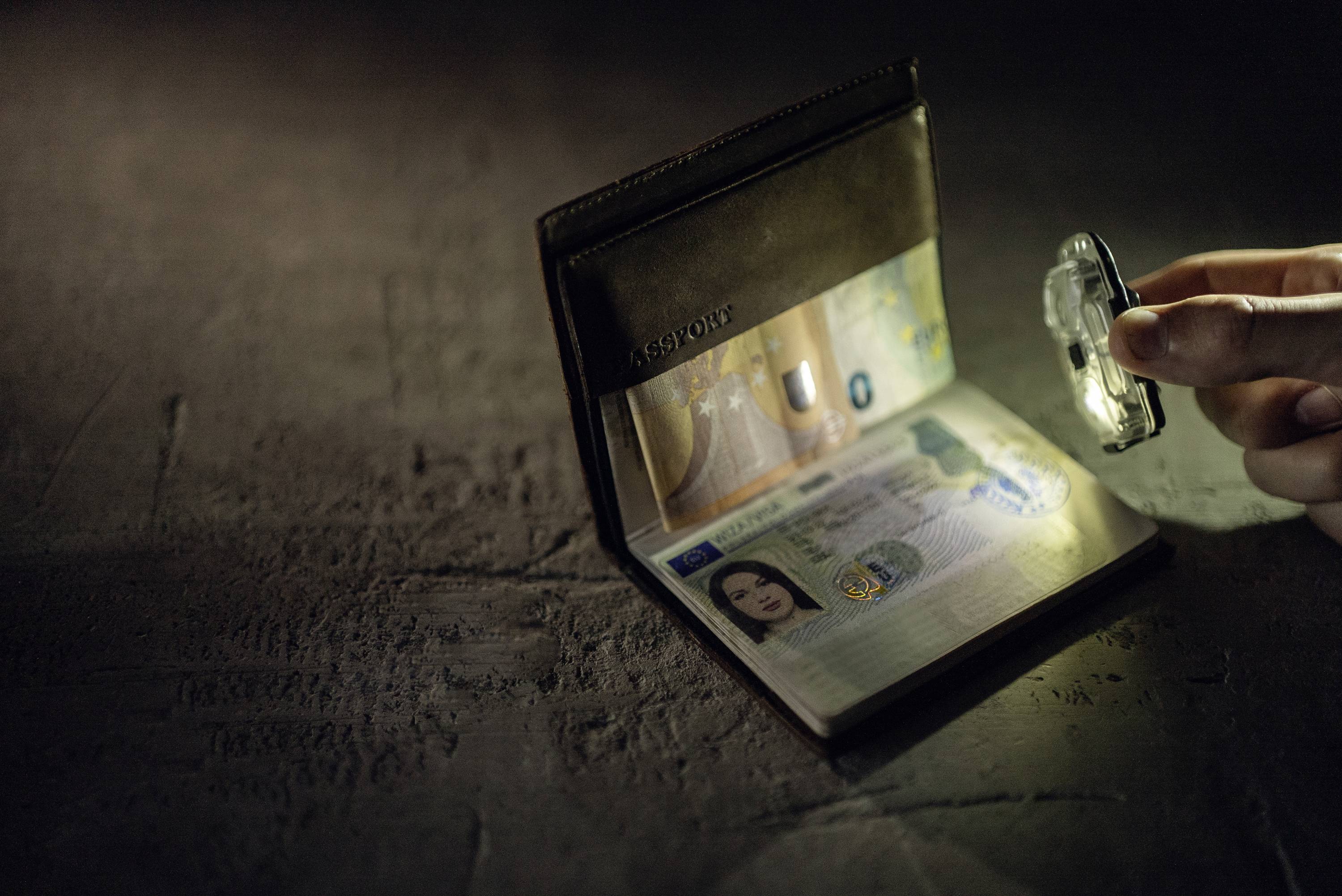 Ein geöffneter Reisepass liegt auf einem Tisch, neben einem Finger, der eine Linse hält. Man sieht das Foto und Sicherheitsmerkmale.