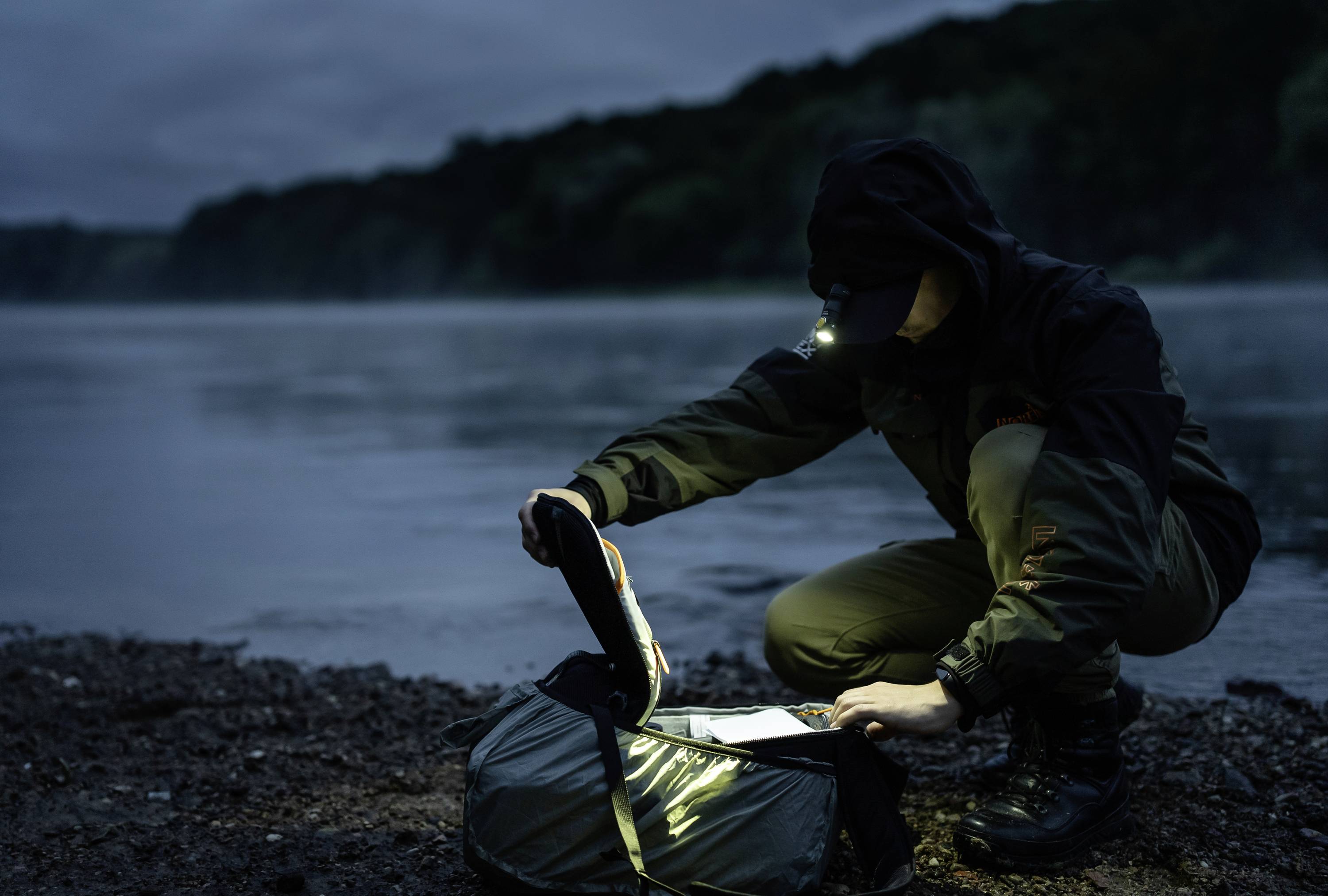 Person am Flussufer, kniend mit Kopflampe, durchsucht eine Tasche bei Dämmerung. Umgeben von Wald, wirkt nachdenklich und konzentriert.