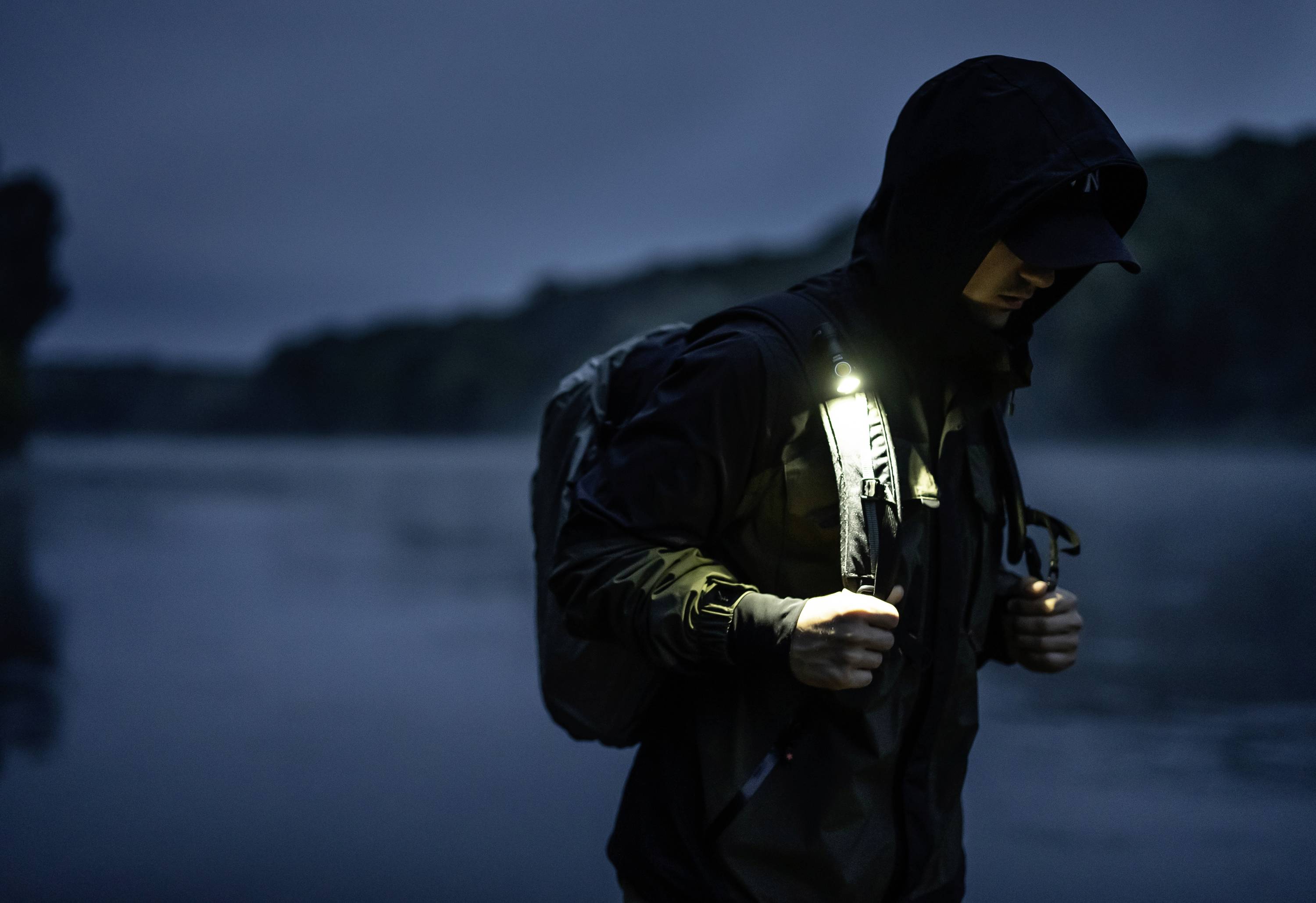 Eine Person wandert bei Dämmerung entlang eines ruhigen Sees. Sie trägt eine dunkle Jacke und einen Rucksack, während sie in die Ferne schaut.