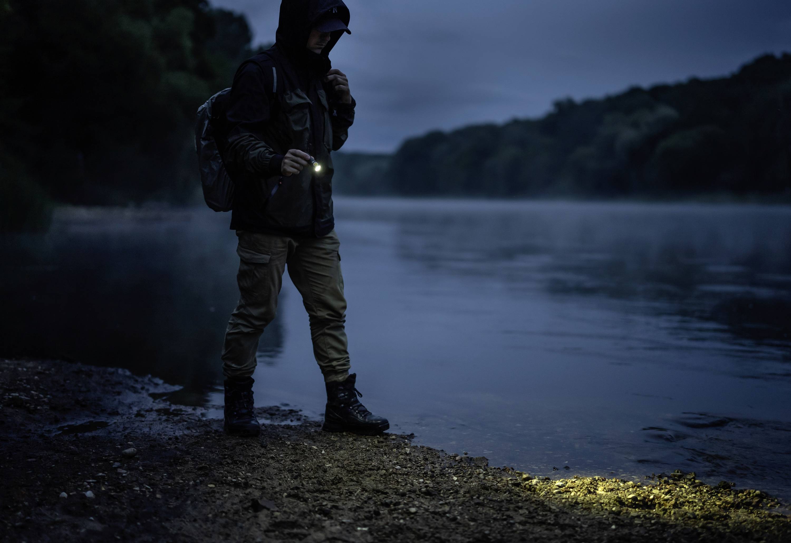 Eine Person mit Kapuze steht nachts am Ufer eines Flusses und leuchtet mit einer Taschenlampe auf den Boden.