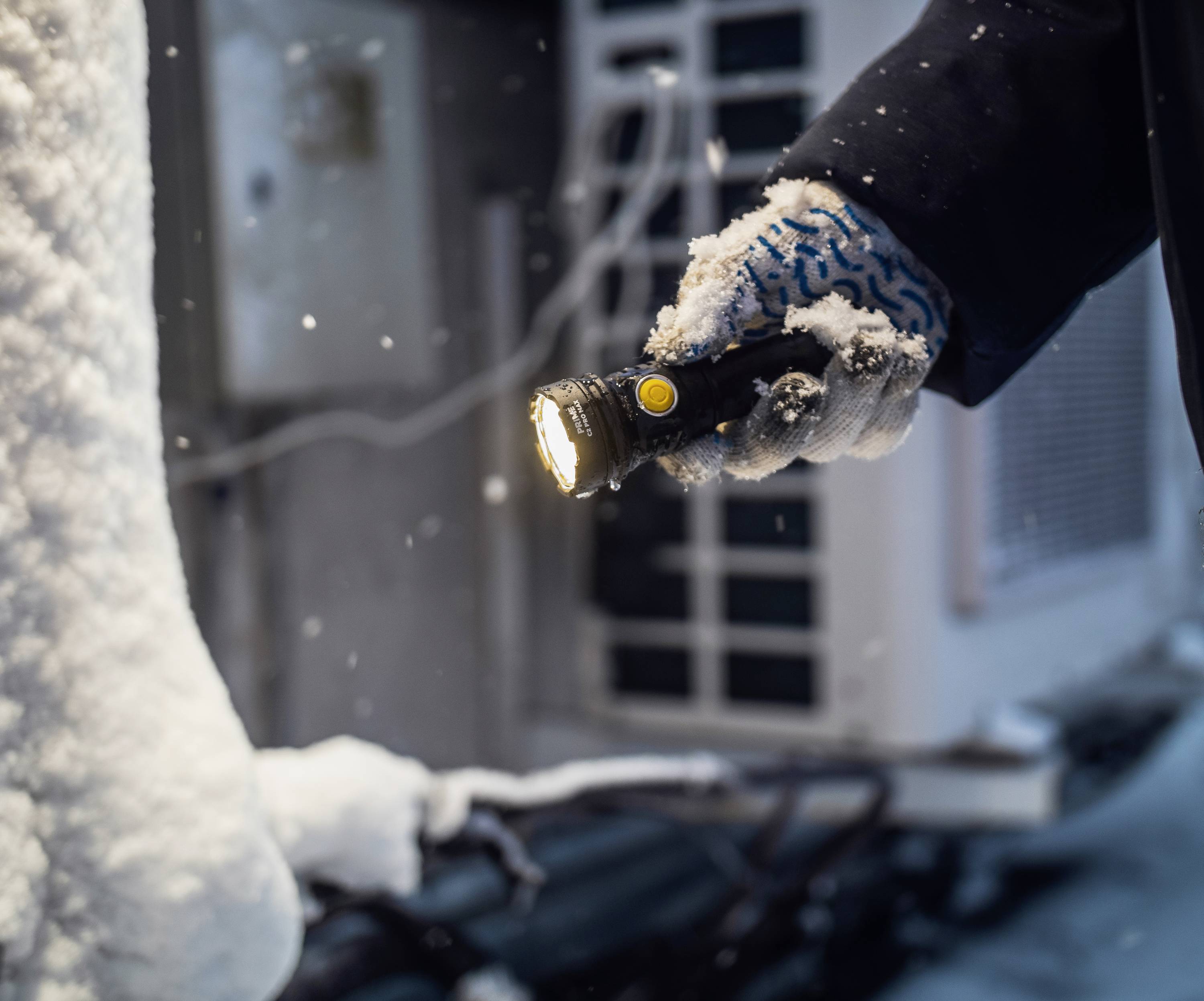 Eine Hand mit Handschuh hält eine leuchtende Taschenlampe im Schnee; dahinter unscharf technisches Gerät im Freien.