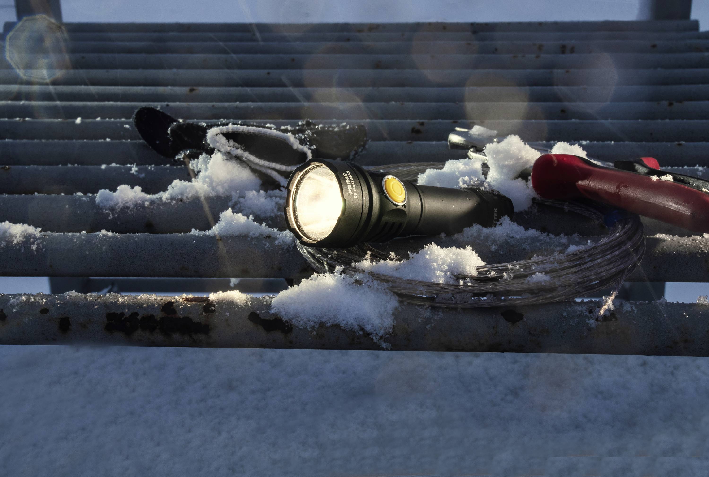 Eine Taschenlampe liegt eingeschaltet auf einem schneebedeckten Gitterrost, umgeben von Werkzeugen und Kabeln bei Tageslicht.