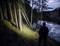 Eine Person steht mit Taschenlampe im Wald am Ufer eines Sees. Es ist Abend; das Wasser reflektiert den wolkigen Himmel.
