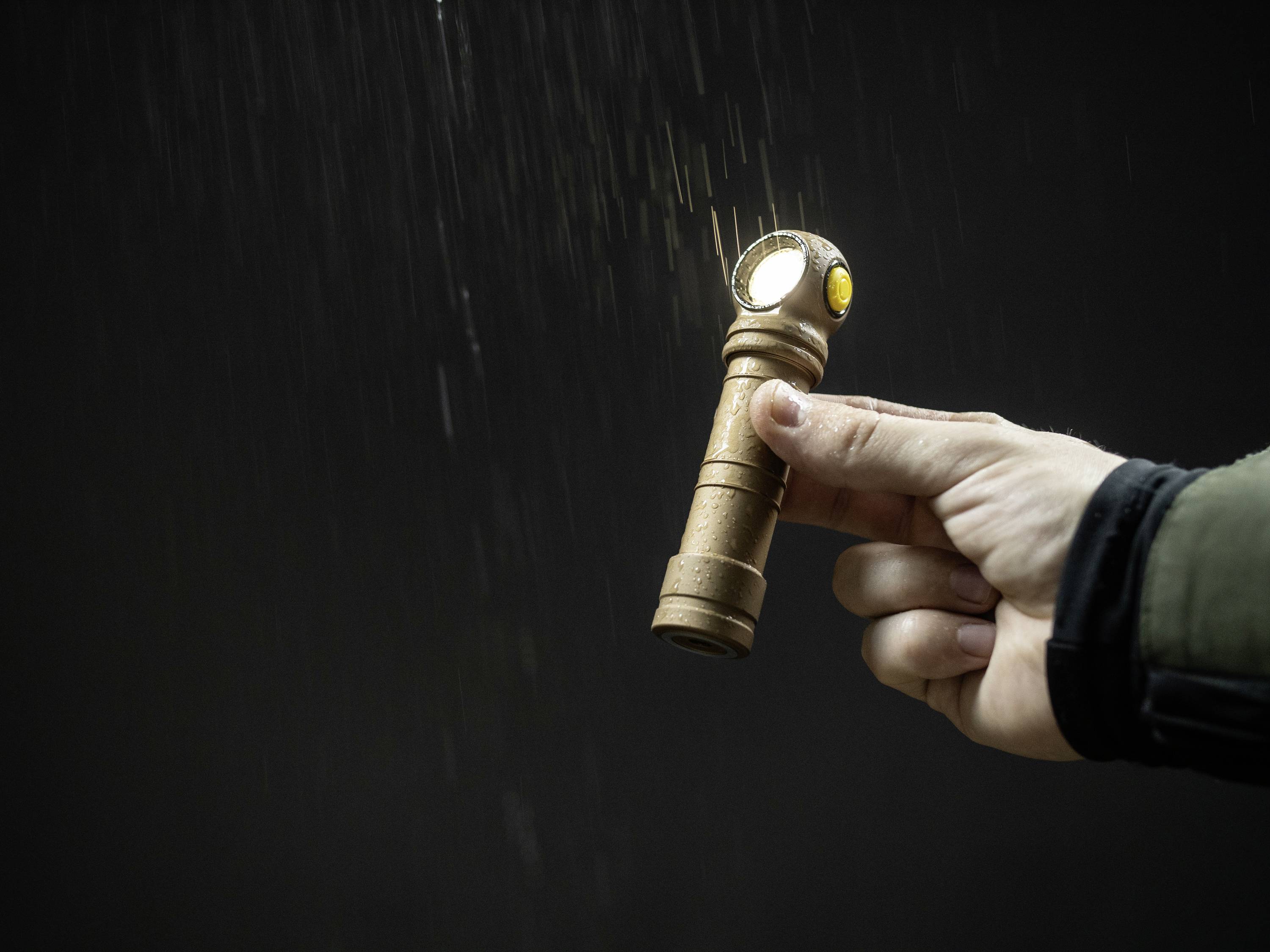 Eine Hand hält eine leuchtende Taschenlampe im Regen. Die Taschenlampe hat einen abgewinkelten Kopf und scheint auf einen dunklen Hintergrund.