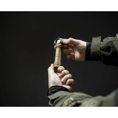 Eine Person hält eine kleine, braune Taschenlampe in der Hand, bereit zum Einsatz. Im Hintergrund ist ein dunkler, neutraler Raum.