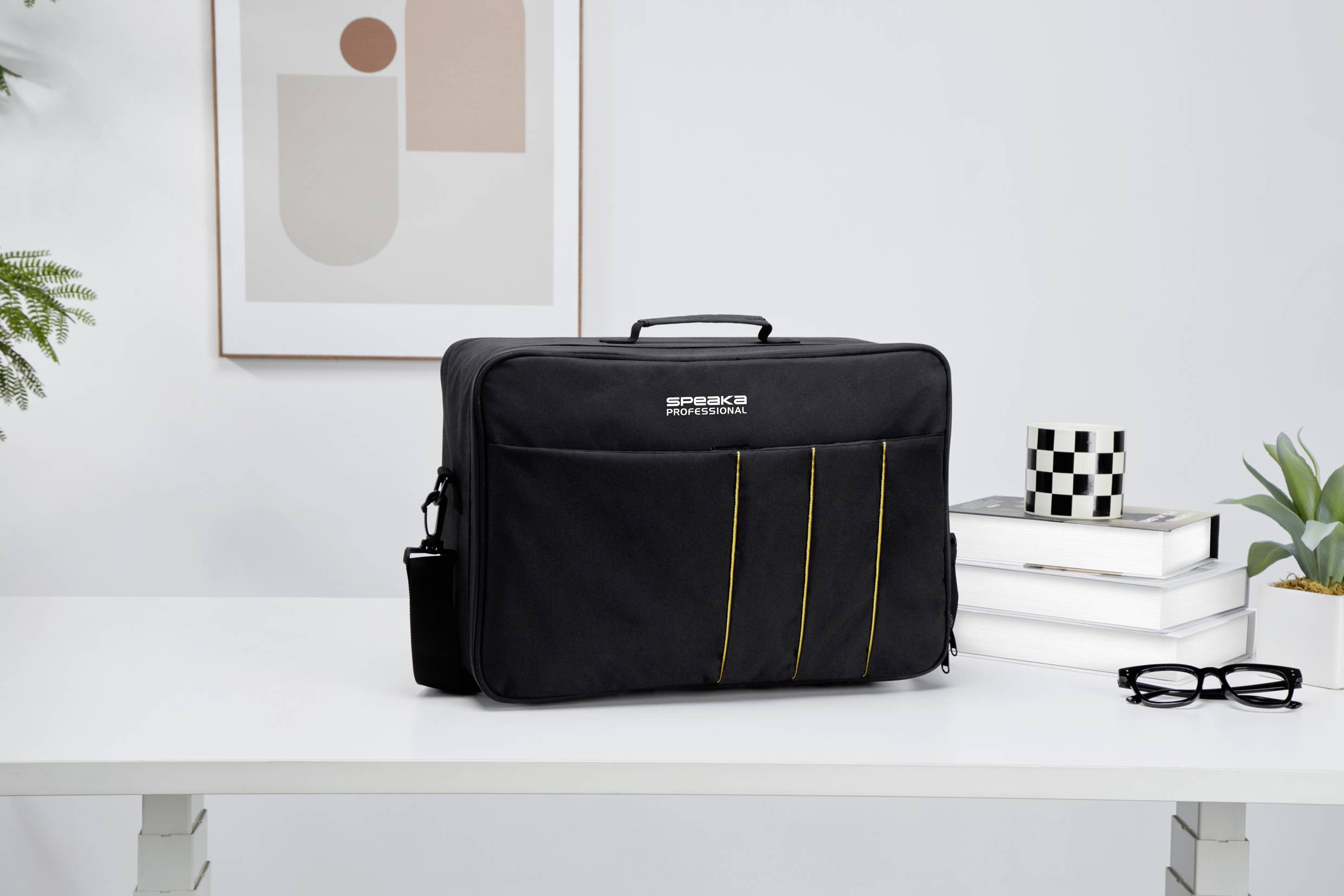 Eine schwarze Laptoptasche steht auf einem weißen Tisch mit Dekorationen, Büchern und einer Brille im Hintergrund.