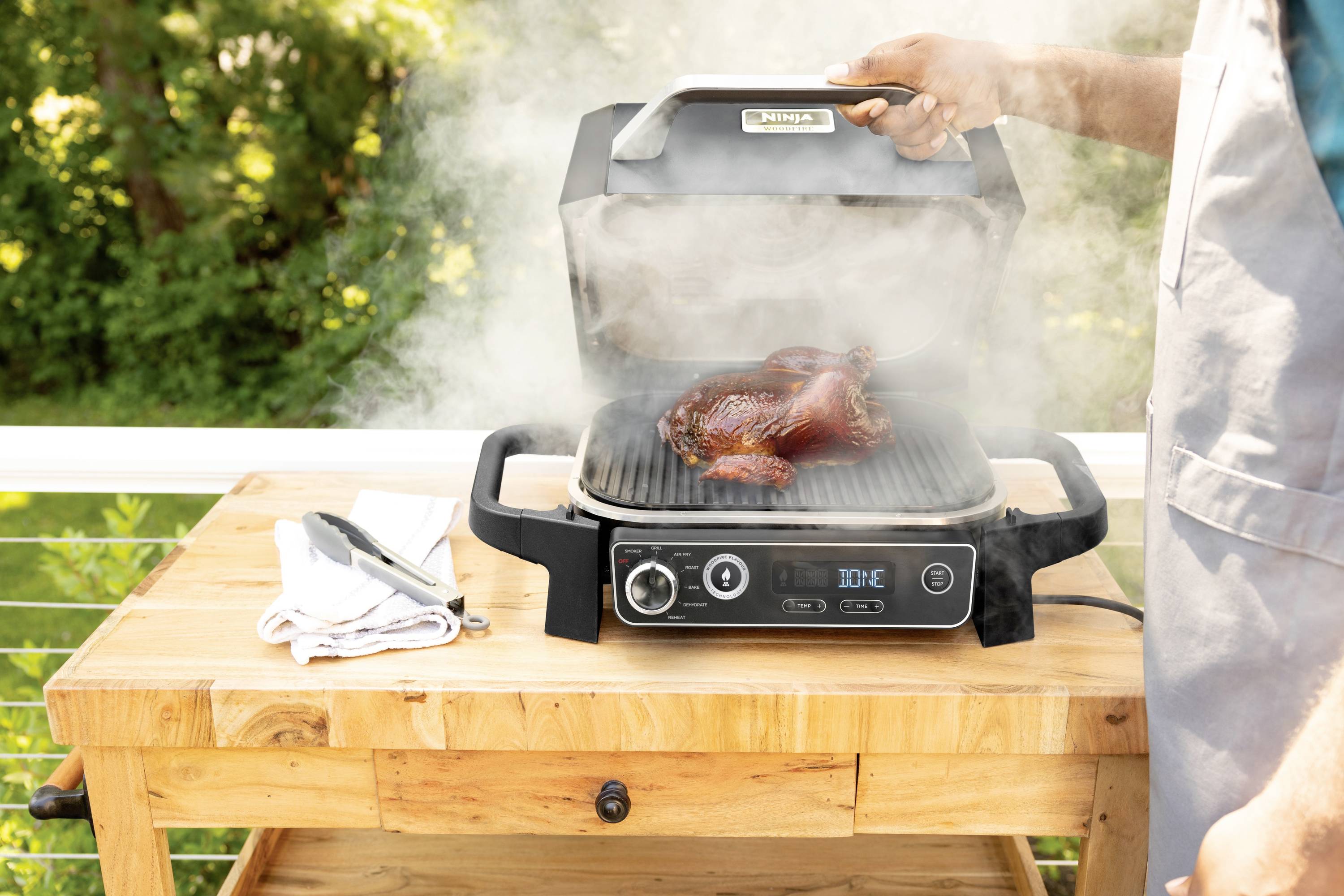 Eine Person hebt den Deckel eines rauchenden Elektrogrills auf einem Holztisch an. Auf dem Grill liegt gegrilltes Fleisch.