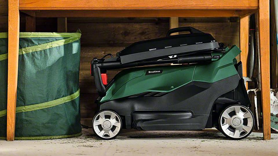 Ein kompakter, grüner Rasenmäher steht zusammengeklappt unter einem Holztisch neben einem großen grünen Sack.