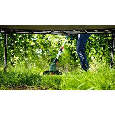 Eine Person mäht mit einem elektrischen Trimmer das hohe Gras unter einem Trampolin. Im Hintergrund sind grüne Büsche sichtbar.