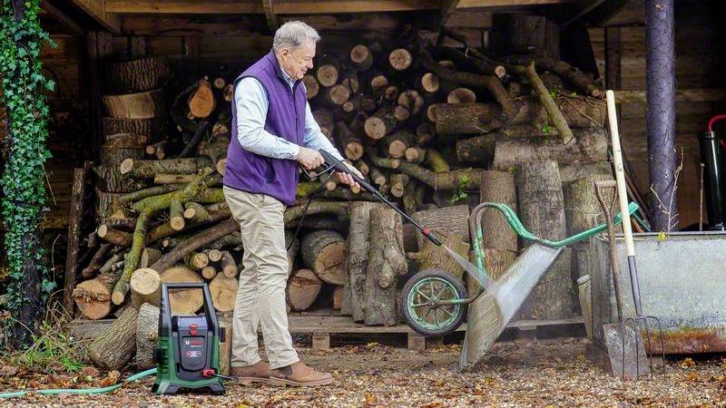 Ein Mann im Freien spritzt mit einem Hochdruckreiniger den Boden sauber. Im Hintergrund sind gestapelte Holzscheite zu sehen.