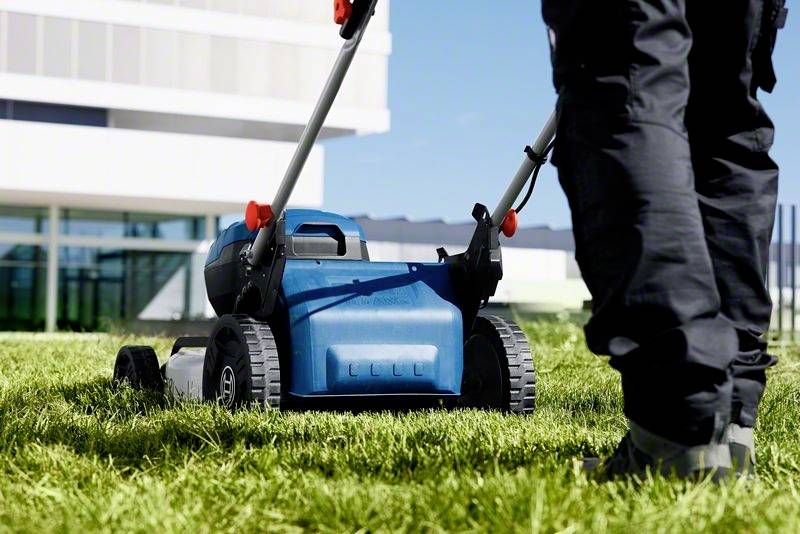 Person mäht Rasen mit einem blauen Rasenmäher bei Sonnenlicht, modernes Gebäude im Hintergrund.
