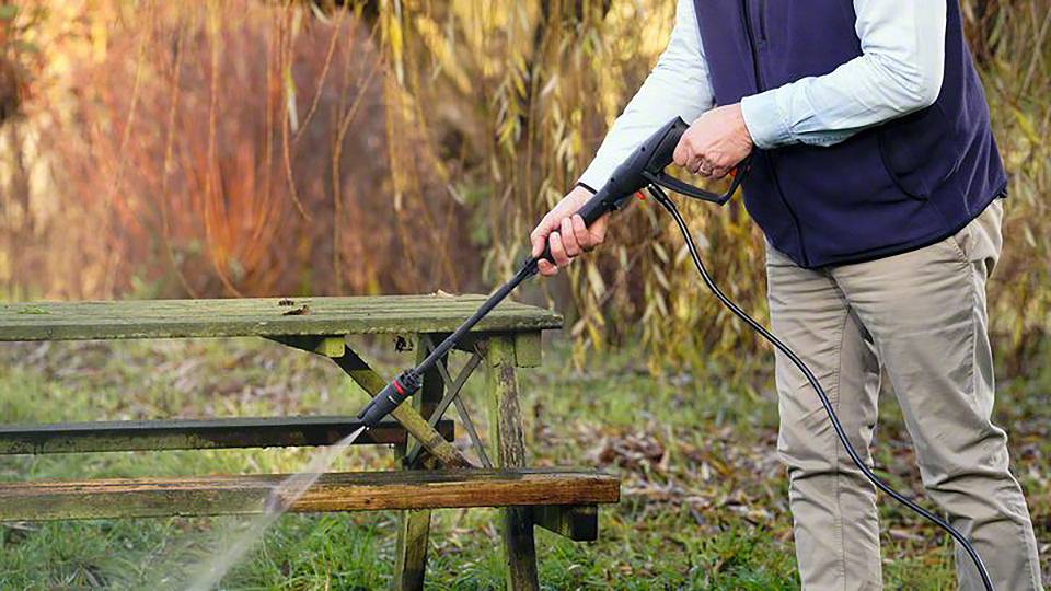 Eine Person reinigt mit einem Hochdruckreiniger eine Holzbank im Freien.