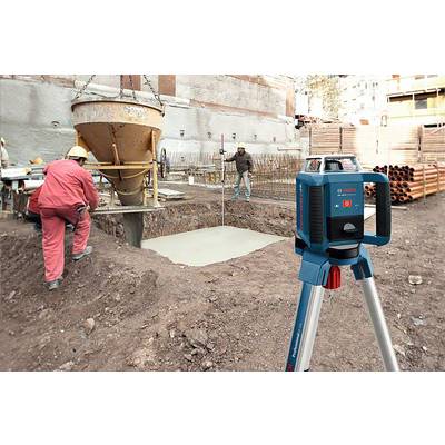 'Bauarbeiter füllen ein Fundamentloch mit Beton auf einer Baustelle, während ein Laser-Nivelliergerät auf einem Stativ ausgerichtet ist.'