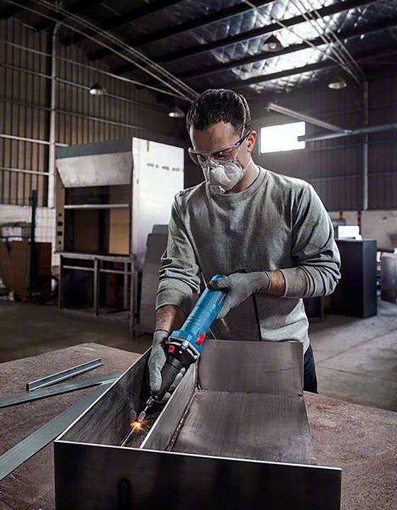 Ein Mann trägt Schutzbrille und Maske und schweißt Metall in einer Werkstatt. Umgeben von Industrieausrüstung und Werkzeugen.