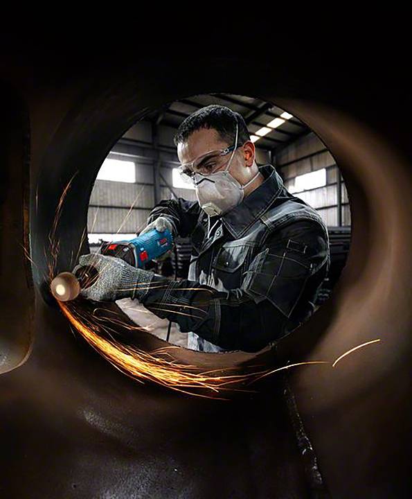 Eine Person trägt Schutzkleidung und schleift mit einem Elektrowerkzeug Metall. Funken fliegen bei der Arbeit in einer Werkstatt.