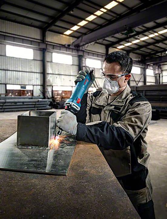 Ein Mann in Schutzkleidung schweißt Metall in einer großen Werkstatt. Funken fliegen, während er ein Werkzeug an einem Metallwürfel verwendet.