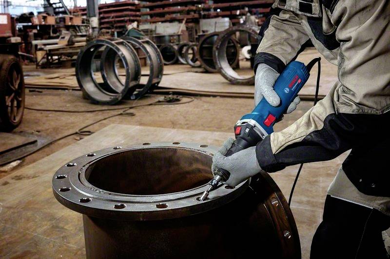 Eine Person in Schutzkleidung bearbeitet eine große Metallflansch-Verbindung mit einem Elektrowerkzeug in einer Werkhalle.