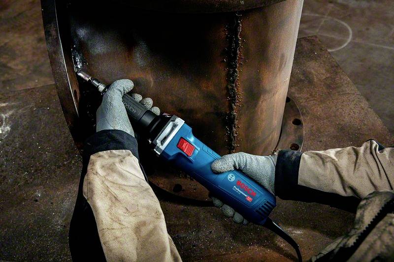 Eine Person schleift mit einem blauen Winkelschleifer an einem großen Metallzylinder. Die Person trägt Handschuhe und Arbeitsschutzkleidung.