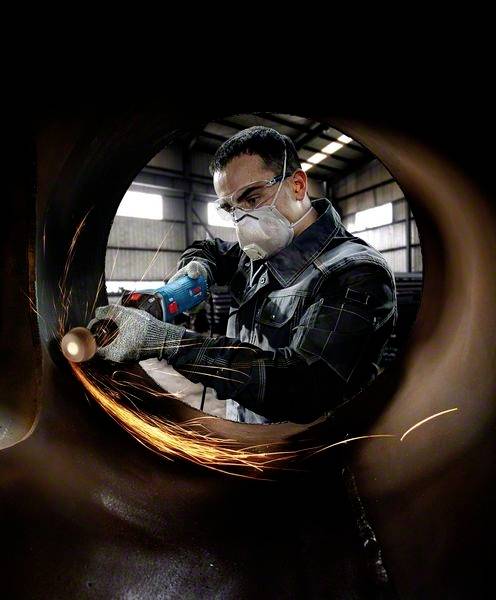 Ein Arbeiter trägt Schutzbrille und Maske, während er mit einem Winkelschleifer an einem Metallstück in einer Werkstatt arbeitet. Funken sprühen.