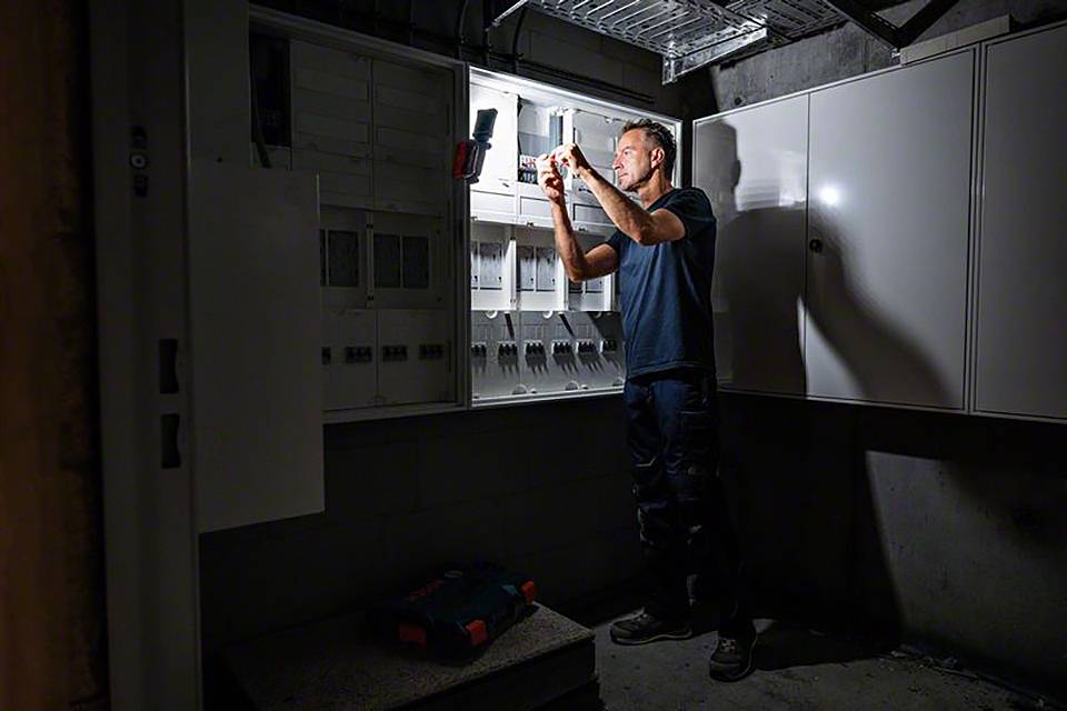 Ein Elektriker untersucht einen offenen Schaltschrank mit einer Taschenlampe. Der Raum ist dunkel, und Werkzeug ist auf dem Boden.