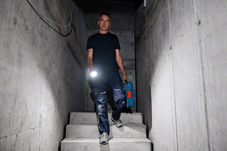 Eine Person steigt mit einer Taschenlampe und einem Werkzeugkoffer eine Betontreppe hinunter, vermutlich in einem Keller.