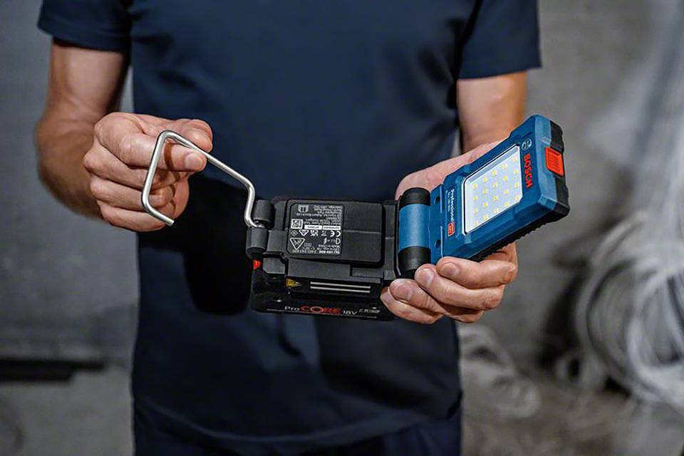 Eine Person hält eine tragbare LED-Arbeitsleuchte und eine Batterie. Die Leuchte hat ein blaues Gehäuse und viele kleine LEDs.
