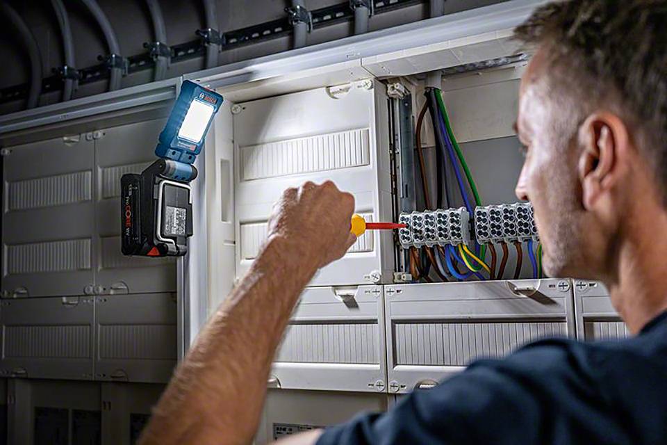 Ein Mann arbeitet mit einem Schraubendreher an einem offenen Schaltschrank voller Kabel. Eine Lampe beleuchtet den Arbeitsbereich.