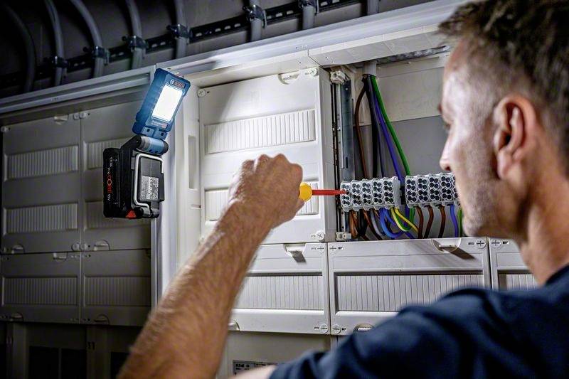 Ein Elektriker arbeitet in einem Schaltkasten, dreht Schrauben an einem Kabelblock. Eine tragbare Lampe beleuchtet den Arbeitsbereich.