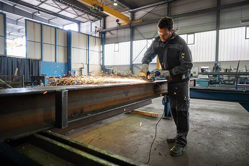 Ein Arbeiter in Schutzkleidung schleift mit einem Winkelschleifer an einem großen Metallträger in einer gut beleuchteten Werkhalle.